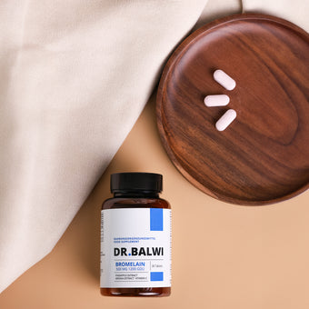 Ein Fläschchen Dr. Balwi Shop Bromelain ruht auf beigem Stoff neben einem Holzteller mit drei rosa Tabletten, die die Vorteile von Ananas Extrakt für alle, die von Haarverlust betroffen sind, aufzeigen.