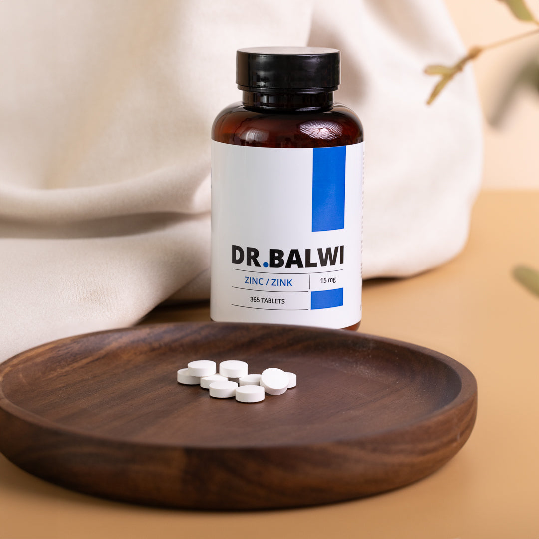 Eine Flasche mit der Aufschrift "Dr. Balwi Shop Zink Tabletten" steht auf einer beigen Fläche neben einer Holzplatte mit mehreren runden Tabletten, die Immunsystem und Haarwachstum unterstützen, vor einem weichen Stoffhintergrund und verschwommenen Pflanzenblättern.