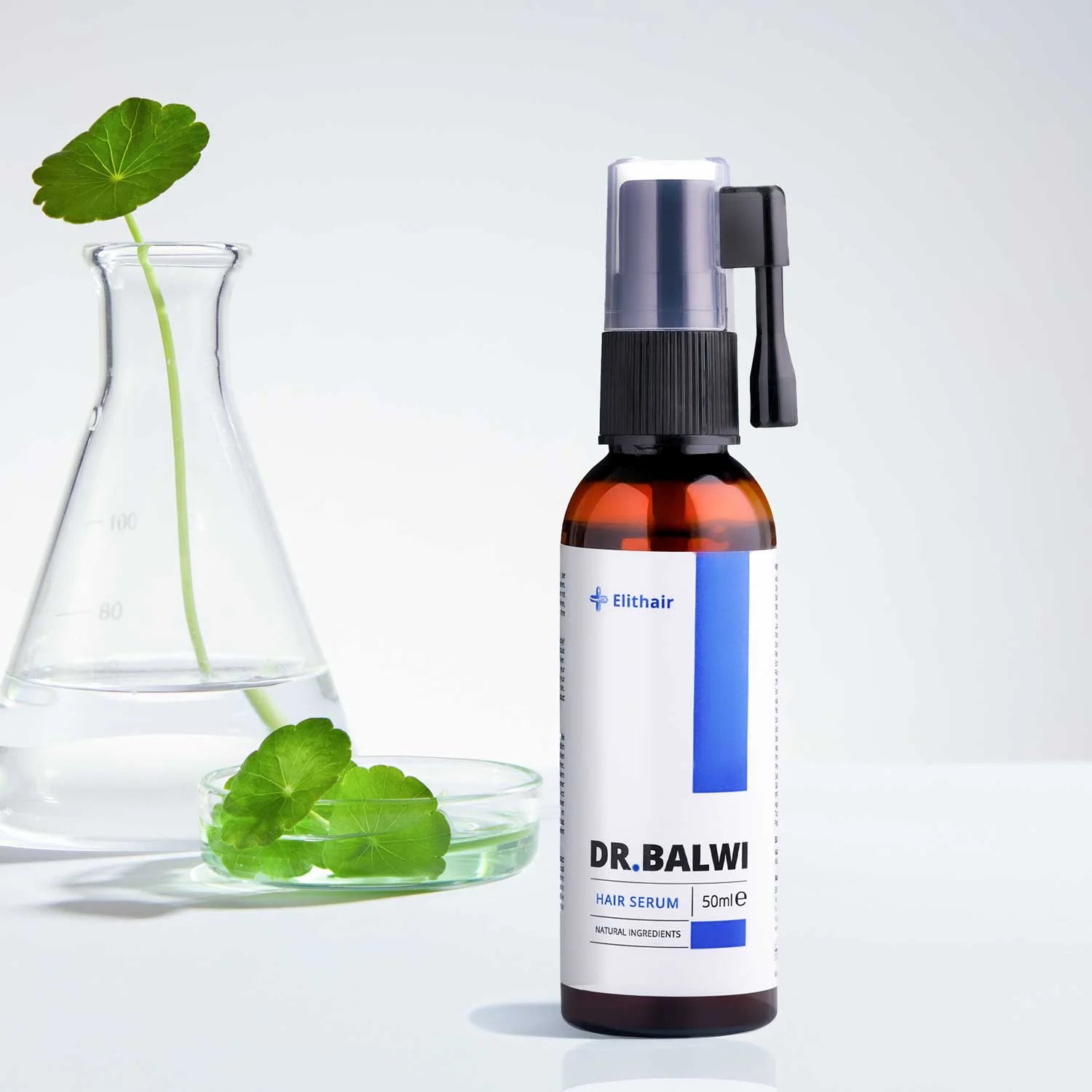 Eine braune Flasche Dr. Balwi Shop Anti Hair Loss Routine mit einer Sprühdüse steht neben einem durchsichtigen Becher und einer Petrischale, die beide grüne Blätter in Wasser enthalten, vor einem hellen Hintergrund.