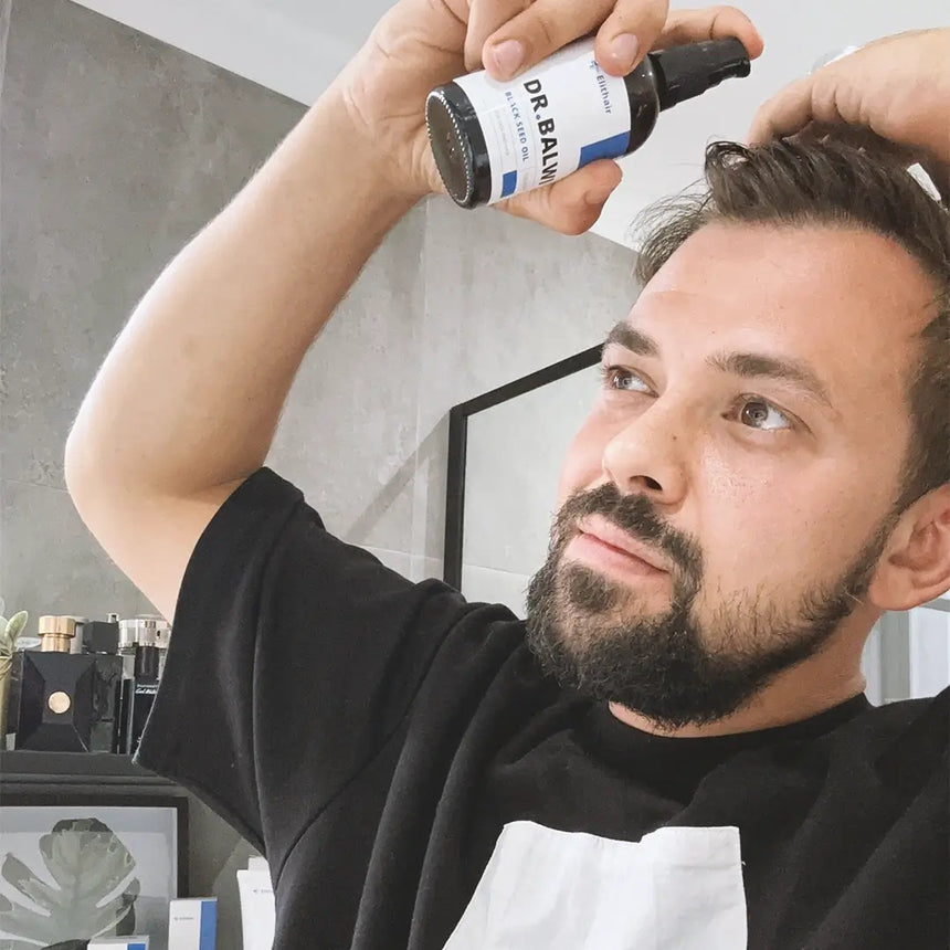 Ein bärtiger Mann in einem schwarzen Hemd trägt in einem modernen Badezimmer mit einer grauen Wand und einem gerahmten Bild im Hintergrund ein Haarprodukt aus einer Sprühflasche mit der Aufschrift DR.BAWL auf sein Haar auf.