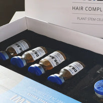 Vier Glasfläschchen mit blauen Kappen, beschriftet mit DR. BAW, befinden sich in einer schwarz ausgekleideten Schachtel. Auf dem Schachteldeckel ist ein Teil des Textes mit der Aufschrift HAIR COMPLEX und PLANT STEM CELL zu sehen.