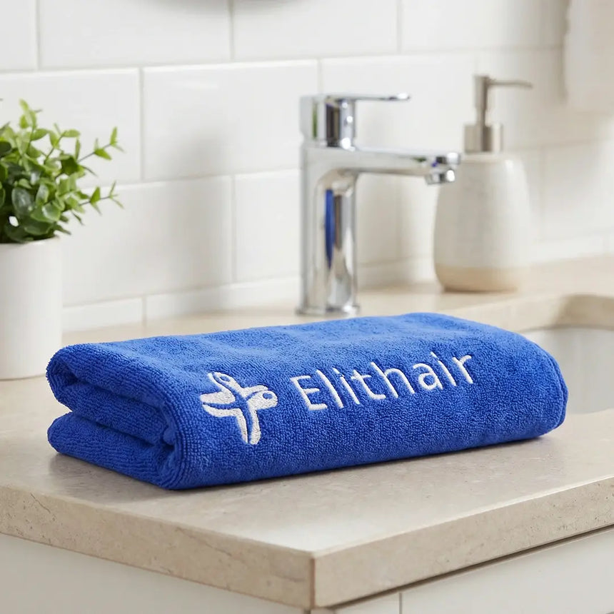 Ein gefaltetes blaues Handtuch mit weißer Elithair-Stickerei liegt auf der Arbeitsplatte eines Badezimmers neben einem Waschbecken, einer Topfpflanze und einem Seifenspender. Im Hintergrund sind weiße Fliesen zu sehen.
