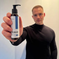 Ein Mann in einem schwarzen Rollkragenpullover hält eine Flasche Dr. Balwi Shampoo in die Kamera, so dass das Produktetikett deutlich sichtbar ist. Der Hintergrund ist unscharf und lenkt die Aufmerksamkeit auf die Shampooflasche.
