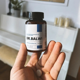 Eine Hand hält eine braune Flasche mit der Aufschrift Biotin & Keratin Tabletten von Dr. Balwi Shop mit einer Tablette auf der Handfläche; ein weißer Tisch und eine unscharfe Wohnungseinrichtung im Hintergrund betonen die Unterstützung für Haarwachstum.