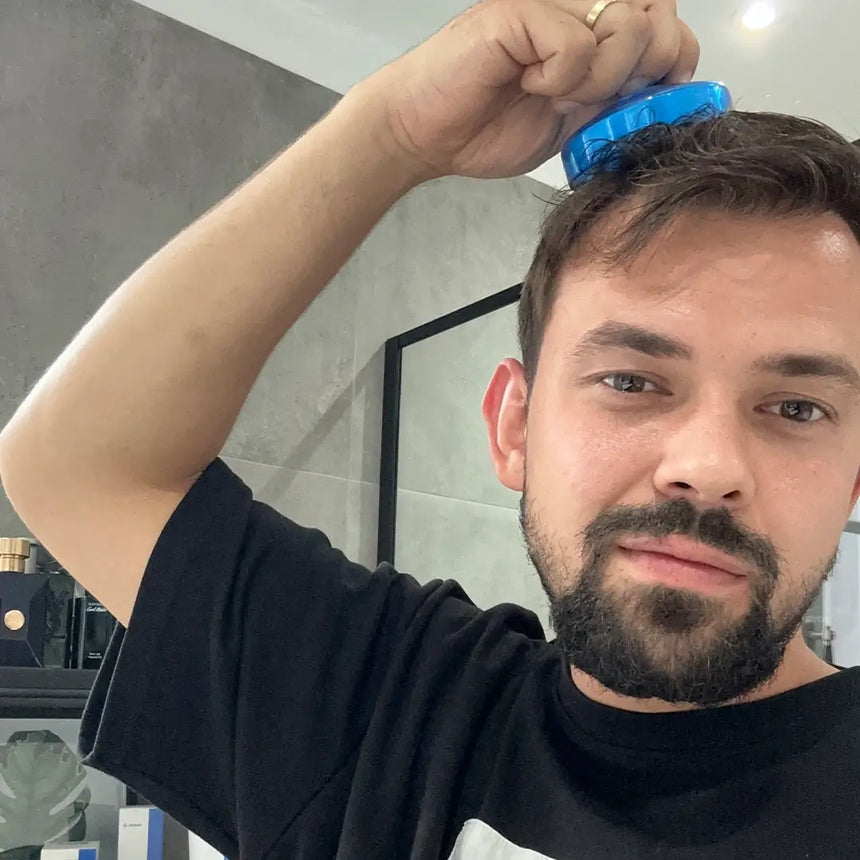 Ein bärtiger Mann genießt eine Kopfmassage mit der blauen Dr. Balwi Massagebürste aus dem Dr. Balwi Shop in einem modernen Badezimmer, vor einem Spiegel und einer grau gekachelten Wand.