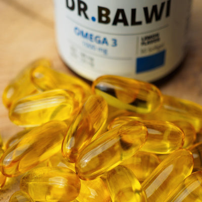 Ein Stapel gelber, durchsichtiger Daily Omega 3 Kapseln sitzt vor einer verschwommenen Dr. Balwi Shop Ergänzungsflasche, die Haarwachstum und gesunde Haarroutinen unterstützt.